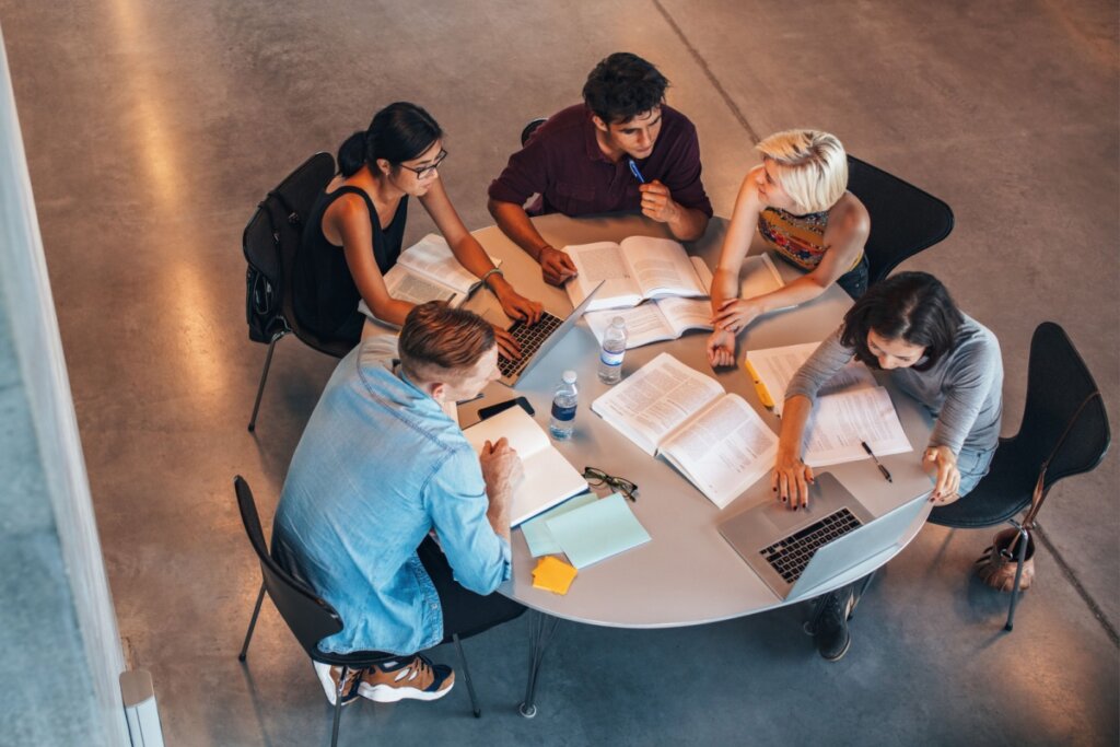 Grupo de jovens sentados reunidos em mesa com livros e notebooks