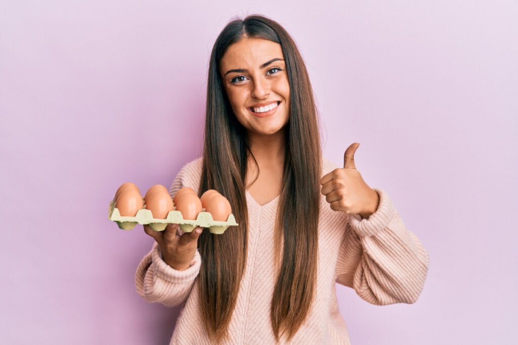 Mulher com o cabelo liso, longo e solto, usando uma camisa de manga longa rosa com uma bandeja de ovo na mão e fazendo um sinal de positivo com a outra em fundo roxo