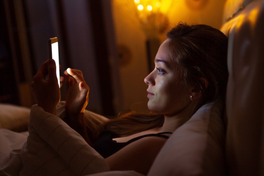 Mulher deitada na cama com o celular na mão e luz da tela refletindo no rosto