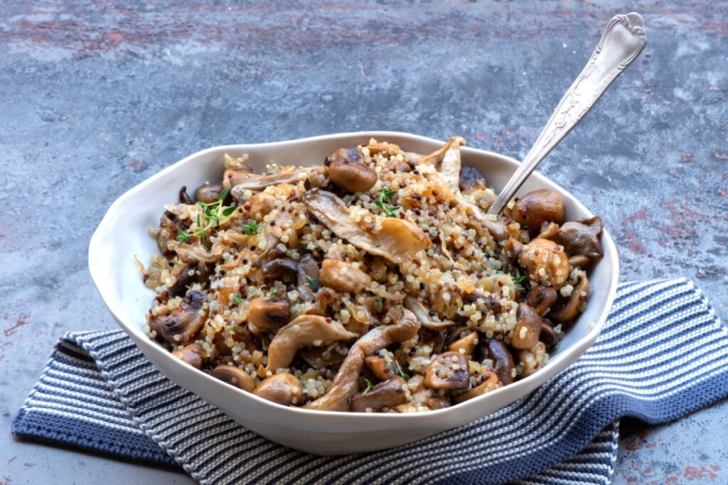 Risoto de quinoa com cogumelo servido em tigela funda branca em cima de pano de prato listrado de branco e azul em cima de bancada de pedra