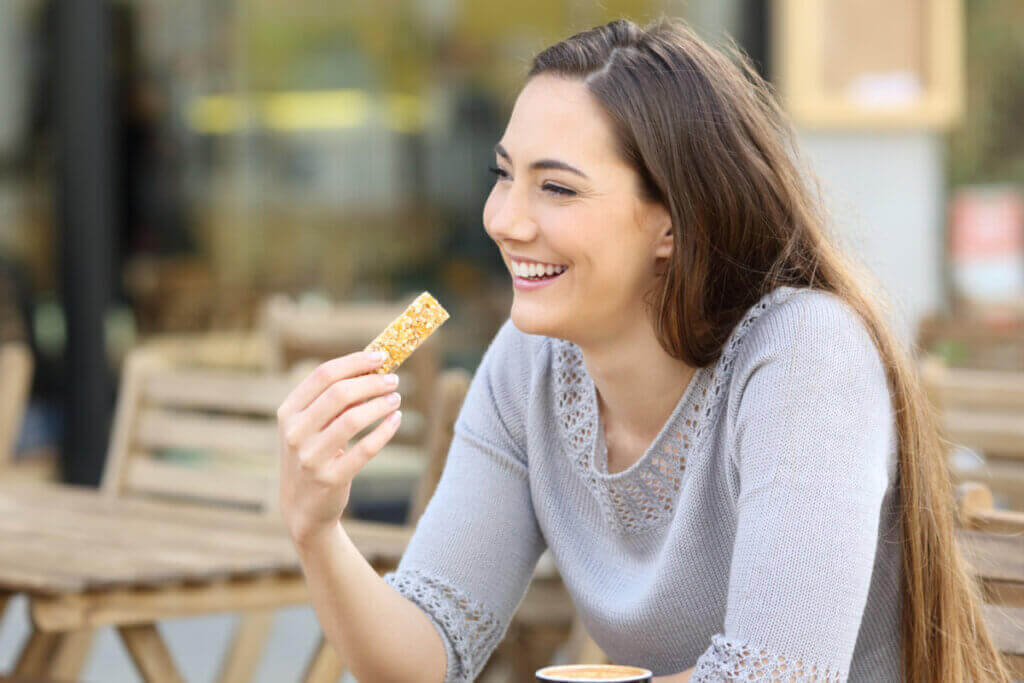 Mulher com cabelo solto, liso e castanho-claro, usando blusa cinza com barra de cereal na mão sentada em mesa ao ar livre