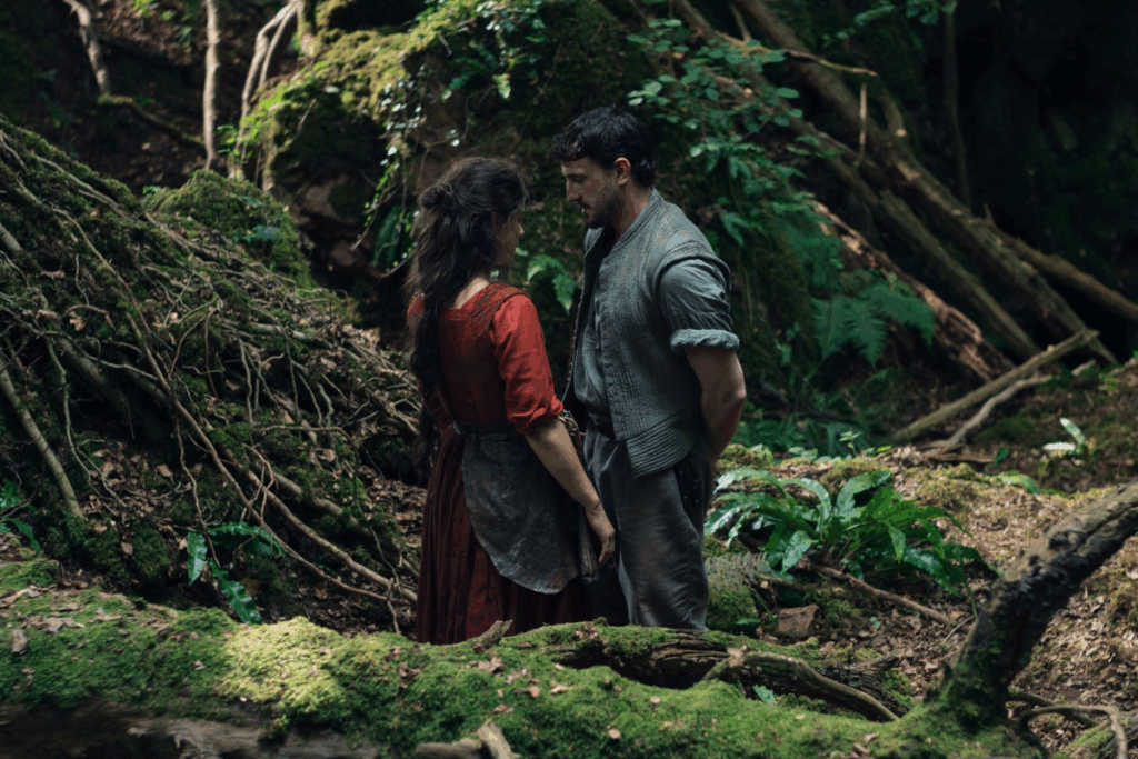Personagens homem e mulher vestidos com trajes de época frente a frente em meio a uma floresta densa, cercados por árvores, raízes e vegetação verde, em clima intimista