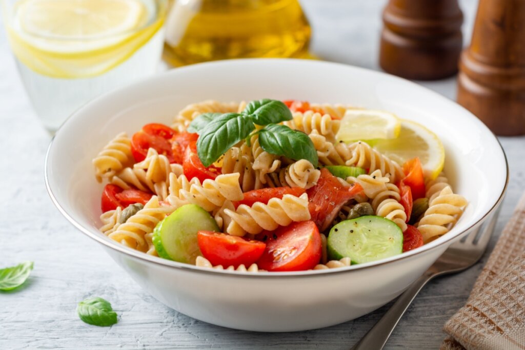 Bowl branco com macarrão integral, tomate-cereja, pepino, azeitona, pedaços de limão-siciliano e folhas de manjericão 