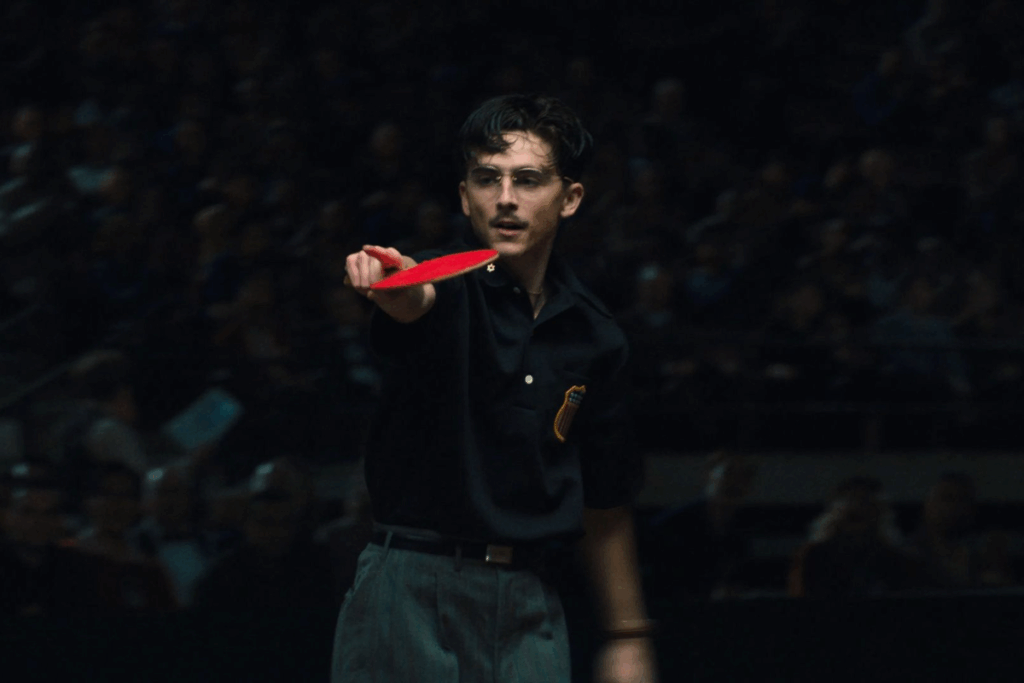 Personagem de Timothée Chalamet segurando uma raquete de tênis de mesa em posição de ataque durante uma partida, em um ambiente fechado, com plateia desfocada ao fundo