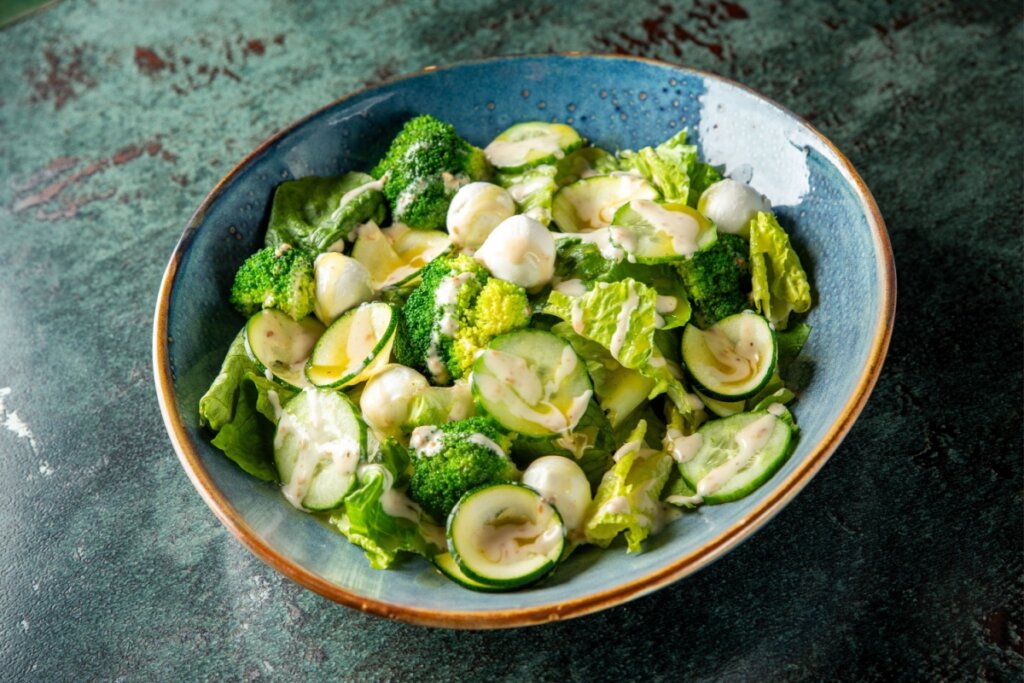 Prato fundo azul com salada com brócolis, muçarela de búfala e pepino regada com molho amarelo e branco 