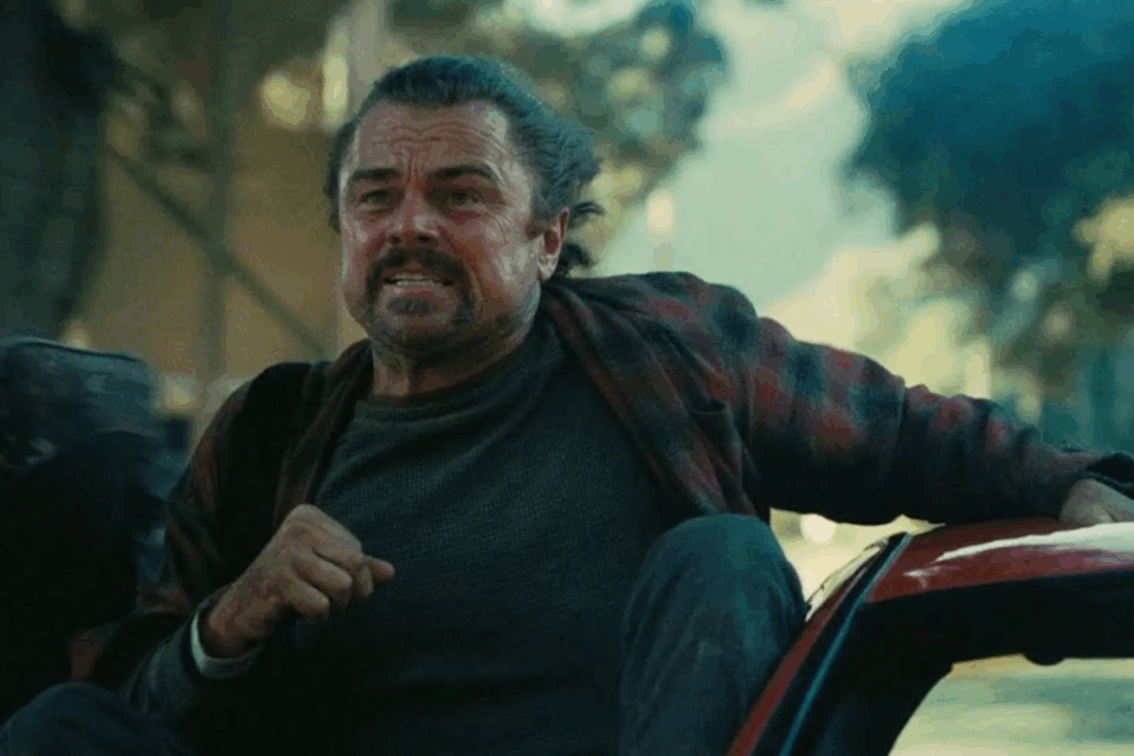 Personagem de Leonardo Di Caprio correndo com expressão de tensão ao lado de um carro, em uma rua arborizada, usando camiseta escura e camisa aberta, em cena de ação ao ar livre