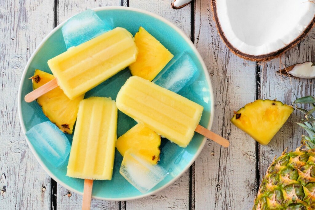 Picolé de abacaxi com coco em prato azul em cima de mesa de madeira com pedaços de gelo e com um coco aberto e um abacaxi ao lado