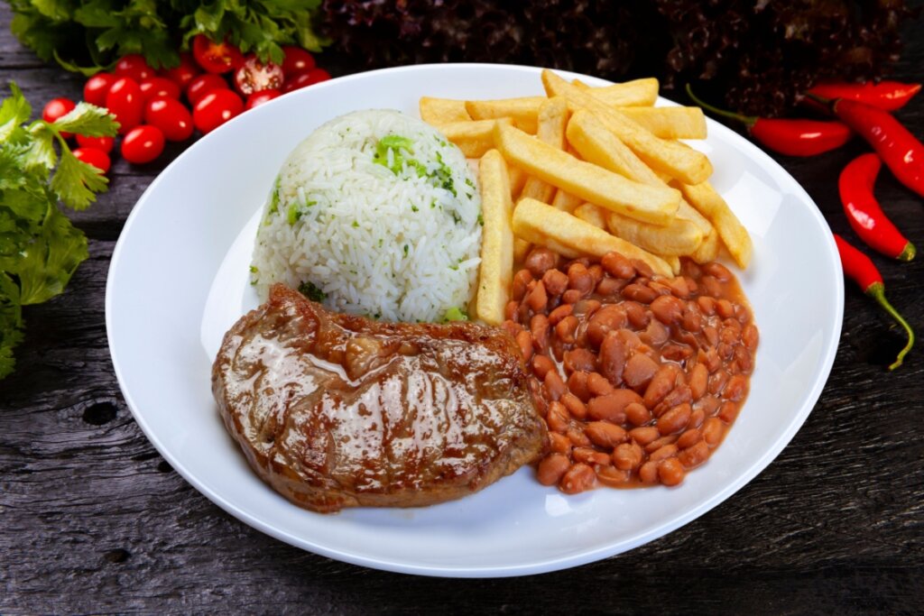 Um prato branco de comida típica brasileira servido sobre uma mesa de madeira escura. O prato contém uma porção de arroz branco com pedaços de brócolis, uma concha de feijão carioca com caldo, um bife grelhado suculento e uma porção de batatas fritas douradas. Ao fundo, para decorar a composição, há alface roxa, mini tomates e pimentas vermelhas frescas.