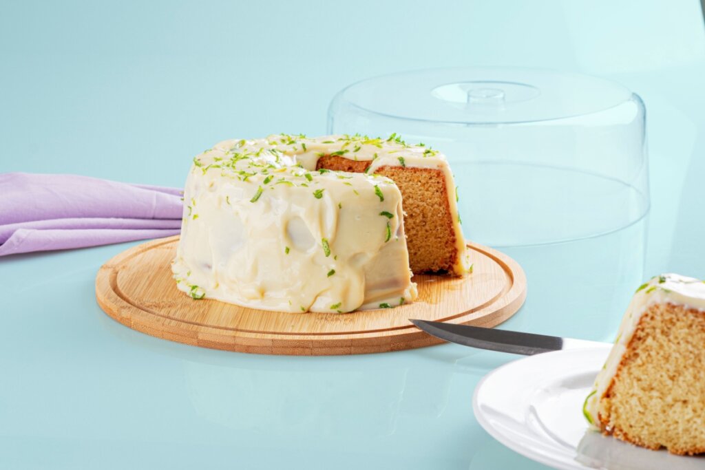 Um bolo de limão caseiro, coberto por uma generosa camada de glacê branco cremoso e raspas de limão verde. O bolo está sobre uma tábua redonda de madeira clara, com uma fatia já cortada e servida em um prato branco em primeiro plano. Ao fundo, uma cúpula de vidro transparente está aberta e há um guardanapo lilás dobrado. A cena é iluminada e limpa, sobre uma superfície azul-claro suave