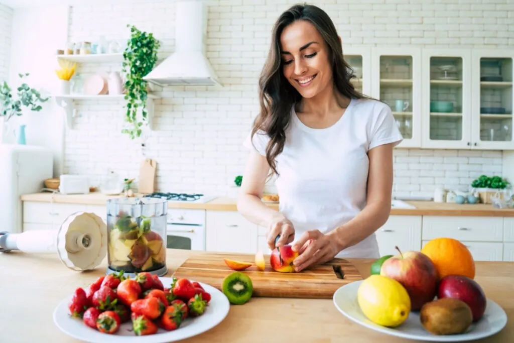 Mulher com cabelo longo, solto, usando camiseta branca cortando frutas em bancada para fazer suco