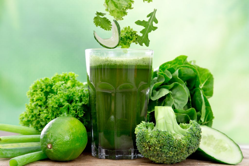 Copo de suco verde-escuro sobre a mesa, cercado por alface crespa, folhas verdes, limão inteiro, talos verdes, brócolis, rúcula, espinafre e fatia de pepino
