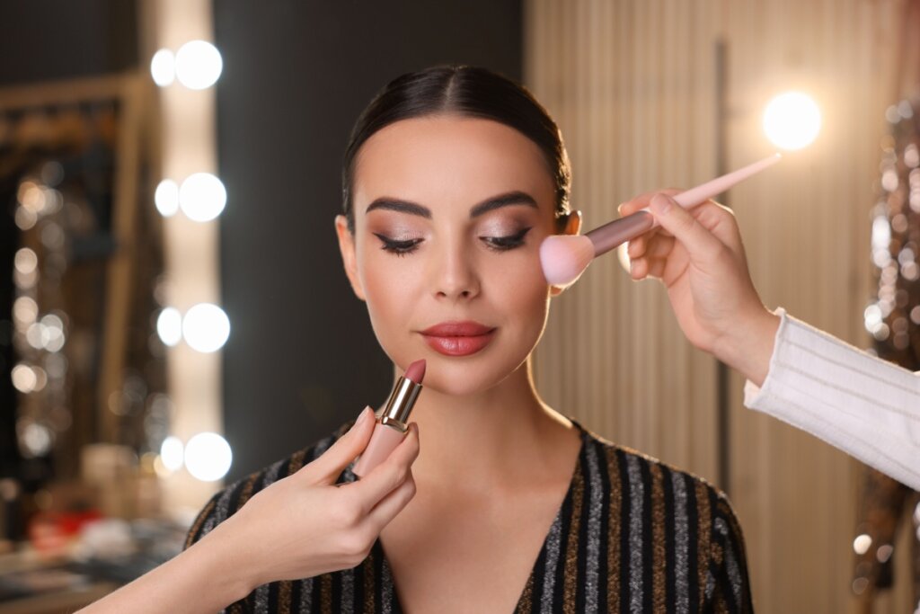 Mãos maquiando o rosto de uma mulher sentada enquanto ela está com os olhos fechados 