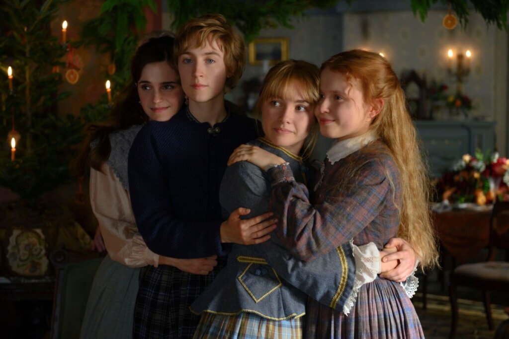 Cena do filme "Adoráveis Mulheres" mostrando as quatro irmãs March — Jo, Meg, Amy e Beth — abraçadas em um momento de carinho e união. Elas vestem roupas de época em tons de azul e marrom. Ao fundo, um ambiente doméstico acolhedor com uma árvore de Natal decorada e velas acesas, criando uma atmosfera quente e emocional.