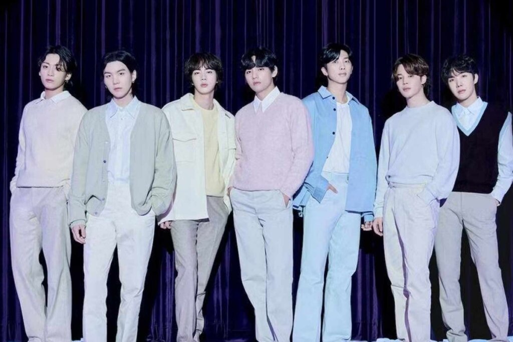 Fotografia de estúdio dos sete membros do grupo sul-coreano BTS. Eles estão alinhados lado a lado à frente de uma cortina azul escura pregueada. Todos vestem roupas em tons pastéis e claros, como branco, bege, azul-claro e rosa, em um estilo casual elegante. A iluminação é suave e destaca os rostos dos integrantes.
