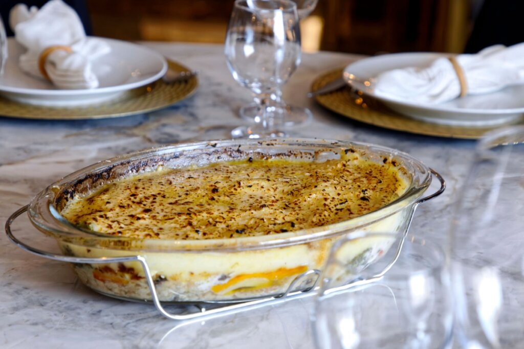 Travessa de bacalhau cremoso gratinado com camada dourada e levemente tostada, servida sobre mesa posta