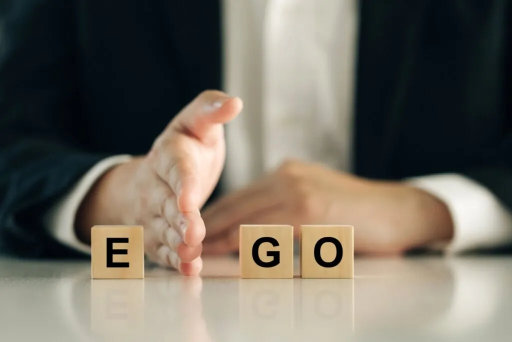 Close das mãos de uma pessoa vestindo paletó escuro, posicionadas atrás de três blocos de madeira que formam a palavra "EGO". Uma das mãos está em pé, como uma barreira, separando a letra "E" das letras "G" e "O". A imagem utiliza um fundo desfocado e tons sóbrios, sugerindo o conceito de controle ou distanciamento do ego.
