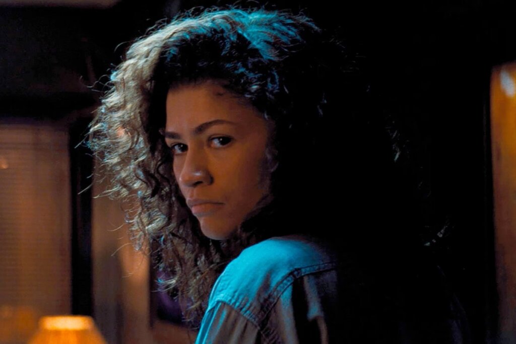 Retrato da atriz Zendaya como a personagem Rue. Ela está de costas, virando o rosto para olhar diretamente para a câmera com uma expressão melancólica e profunda. Seus cabelos são escuros, cacheados e volumosos, iluminados por uma luz azul vinda de cima. O ambiente ao redor está escuro, criando um clima introspectivo e sombrio.