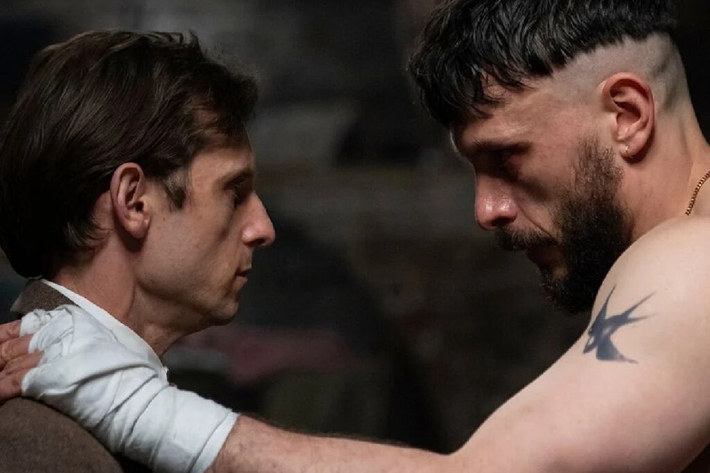 Cena dramática em close-up de dois homens se encarando intensamente. À esquerda, um homem de perfil com expressão séria. À direita, um homem com corte de cabelo curto (estilo "militar" com as laterais raspadas), barba e uma tatuagem de pássaro no ombro. Ele segura o ombro do outro homem, que está com a mão enfaixada com ataduras brancas. A iluminação é baixa e contrastada.
