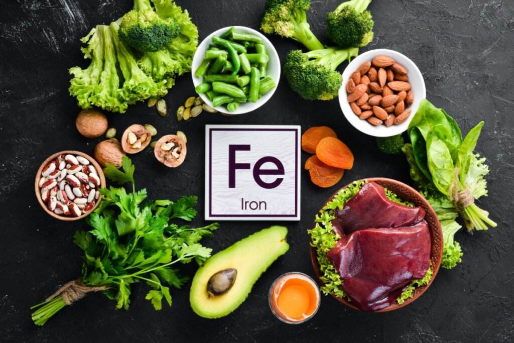 Alimentos ricos em ferro dispostos em cima de mesa de pedra, como carne vermelha, abacate, leguminosas e vegetais verdes e com uma plaquinha com o símbolo do ferro