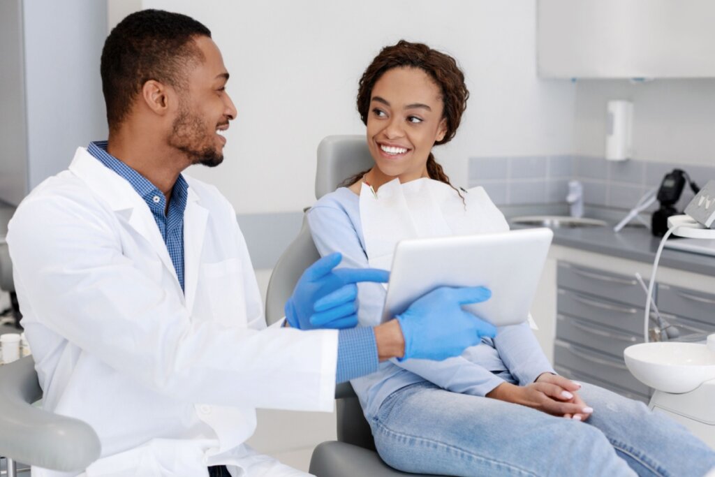 Jovem com cabelo cacheado preso usando casaco azul e calça jeans em consulta com dentista que está sentado ao lado, tem o cabelo curto, barba, esta usando camisa azul e jaleco branco mostrando algo em tablet