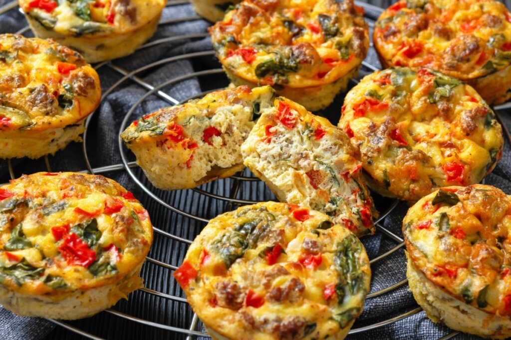 A imagem mostra pequenos muffins salgados dispostos sobre uma grade. Eles têm aparência dourada e são recheados com ingredientes como vegetais e ervas