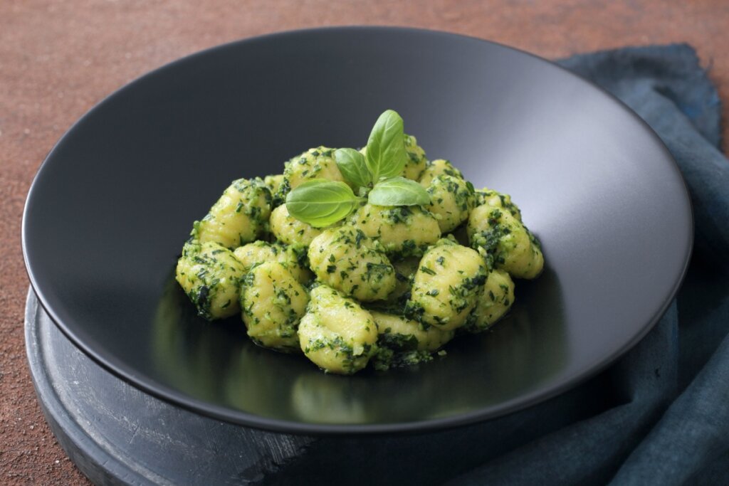 Nhoque ao molho pesto servido em prato fundo preto em cima de mesa de madeira