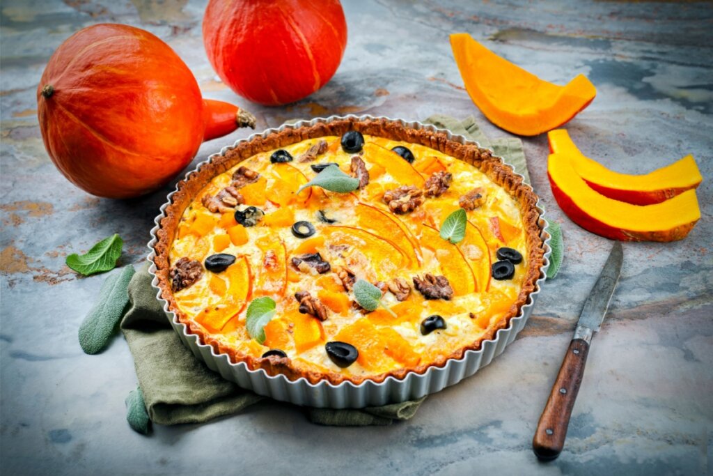 Quiche de abóbora com nozes e azeitonas  em cima de uma mesa cinza com pedaços de abóbora