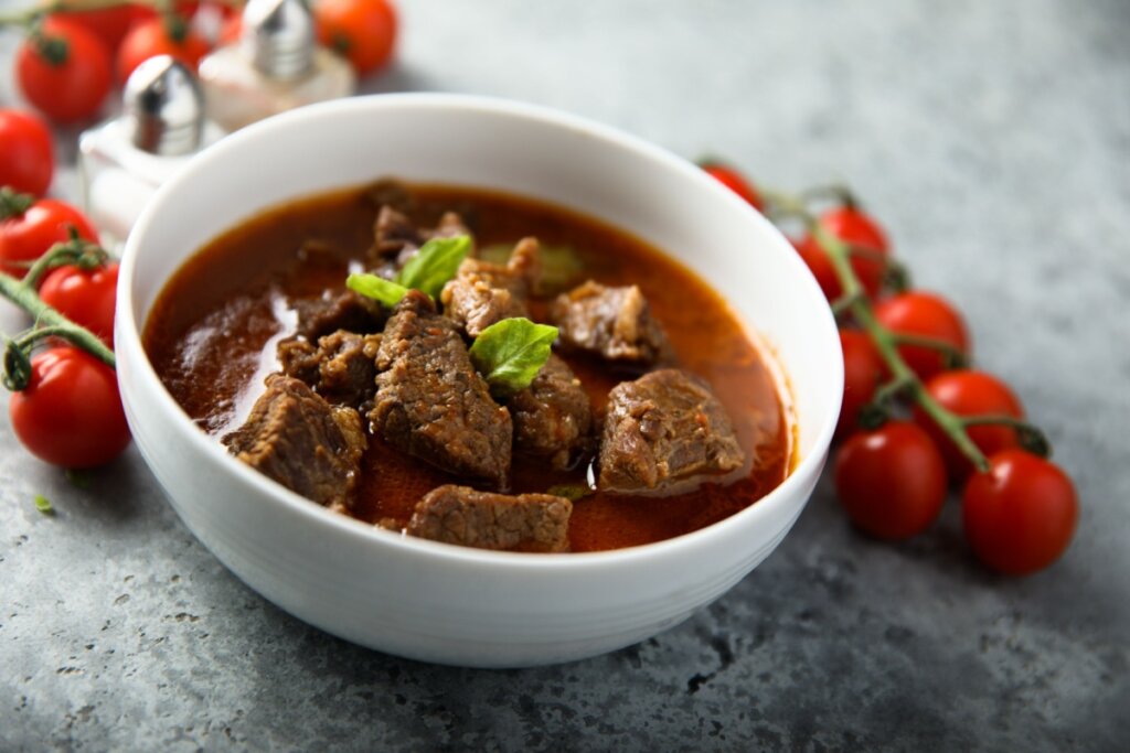 Uma tigela branca de cerâmica contendo um ensopado de carne bovina em cubos, imersos em um molho denso e avermelhado. Sobre a carne, há pequenas folhas de manjericão fresco para decorar. Ao fundo, sobre uma superfície cinza marmorizada, aparecem ramos de tomates-cereja bem vermelhos e pequenos saleiros de vidro. A iluminação foca no centro da tigela, destacando a suculência da carne.