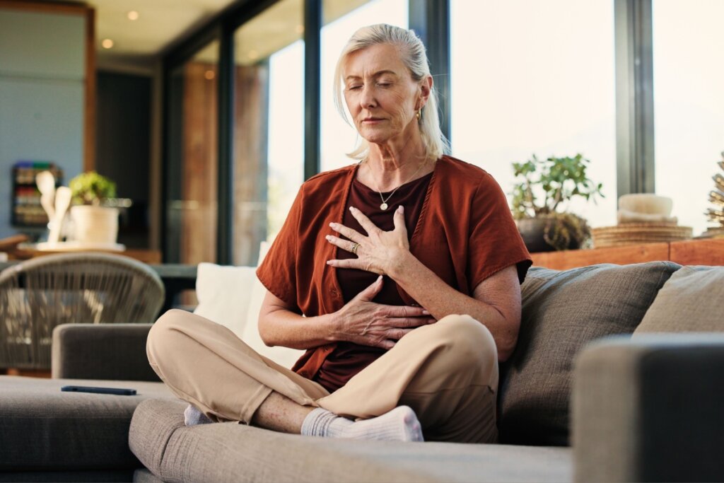 Uma mulher madura com cabelos loiros está sentada em posição de lótus sobre um sofá cinza em uma sala de estar ampla e iluminada. Ela está com os olhos fechados, uma expressão serena, e mantém as mãos espalmadas sobre o peito e o abdômen, sugerindo uma prática de meditação ou respiração consciente.