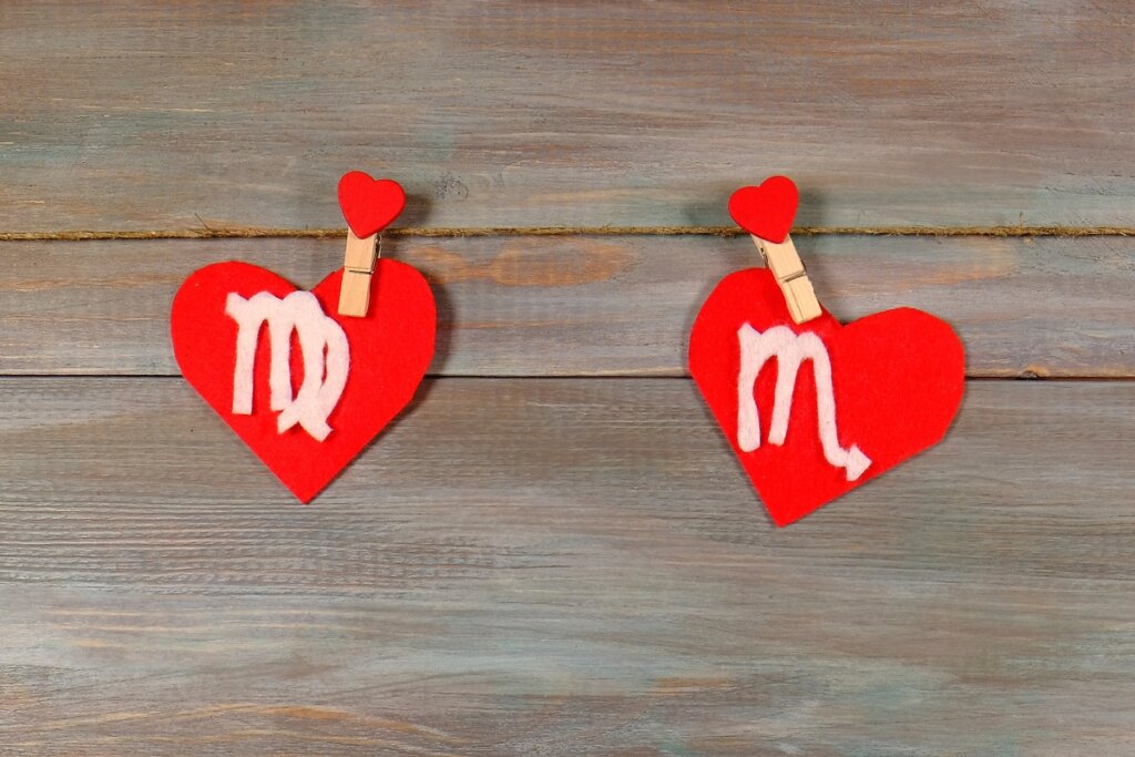 Dois corações de feltro vermelho pendurados em um varal de corda natural sobre um fundo de madeira. O coração à esquerda mostra o símbolo de Virgem e o coração à direita mostra o símbolo de Escorpião.