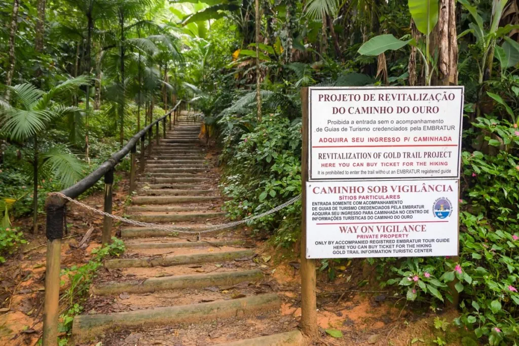 Escadaria no meio da mata com placa informativa sobre o Caminho do Ouro e acesso controlado