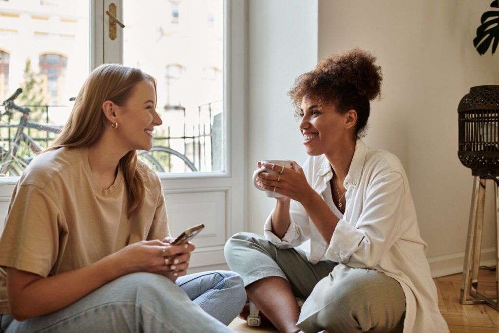 Duas mulheres sentadas em frente a uma janela, segurando xícaras, conversando