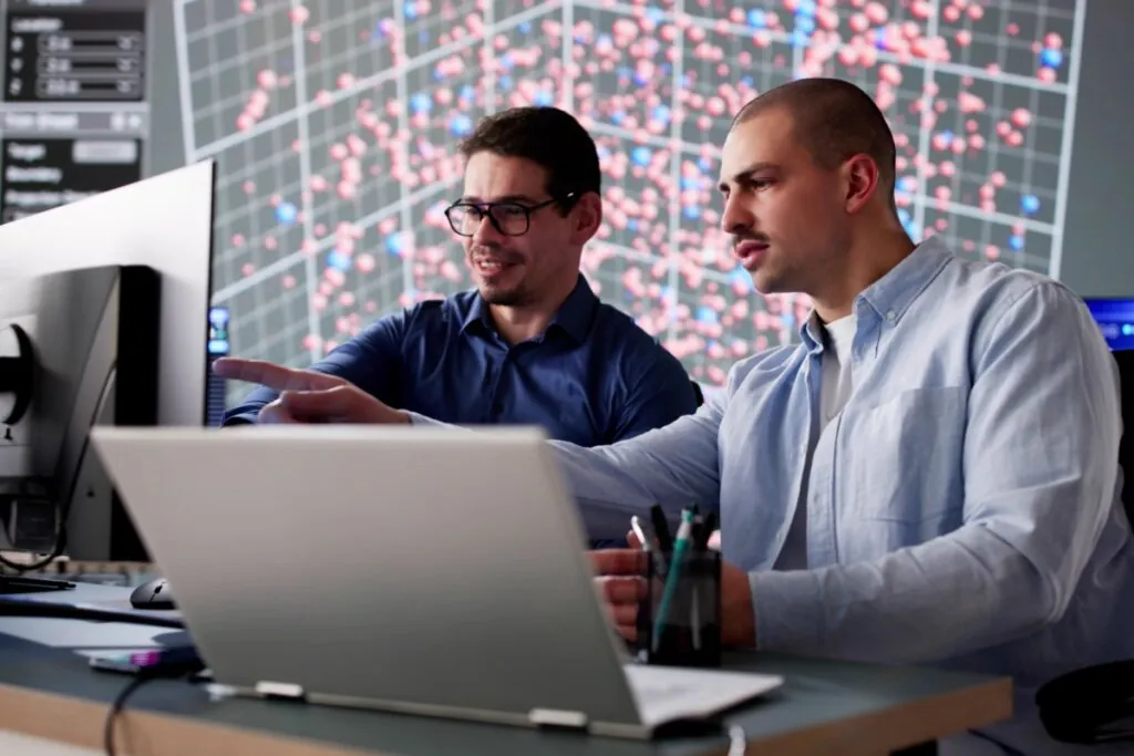 Dois homens trabalham em um ambiente de escritório tecnológico, focado em análise de dados. Um deles, de óculos e camisa social azul escura, aponta para a tela de um monitor de computador enquanto conversa com o colega. O segundo homem, com cabelo curto e camisa social azul clara sobre uma camiseta branca, observa atentamente a tela. Ao fundo, um grande painel digital exibe um gráfico tridimensional complexo com diversos pontos de dados em tons de vermelho e azul sobre uma grade. Na mesa, em primeiro plano, vê-se a parte traseira de um laptop prateado e um porta-canetas.