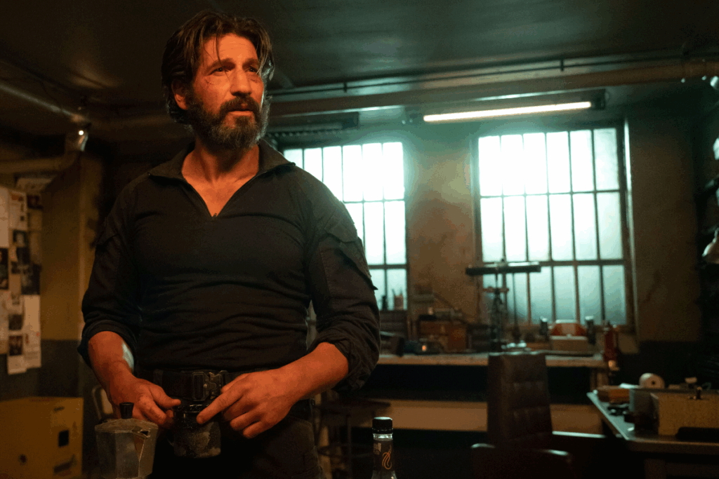 Homem com cabelo grande, barba grande, usando camisa preta de manga longa e calça preta em cozinha escura