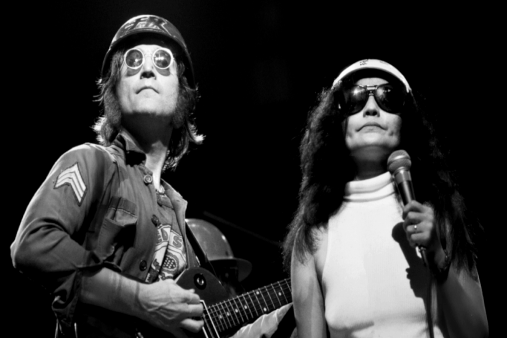 Foto em preto e branco com um homem usando boina, e casaco com guitarra e usando óculos escuro e mulher ao lado com o cabelo solto, usando boina branca e regata de gola alta branca e óculos de sol com um microfone na mão