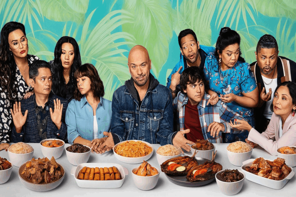 Mesa de jantar com toalha branca com diversos pratos com comida e um grupo de pessoas atrás olhando umas para as outras em fundo azul com plantas em verde-claro