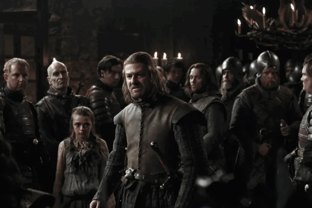 Homem de cabelos perto do ombro diante de um grupo em ambiente medieval na série Game of Thrones