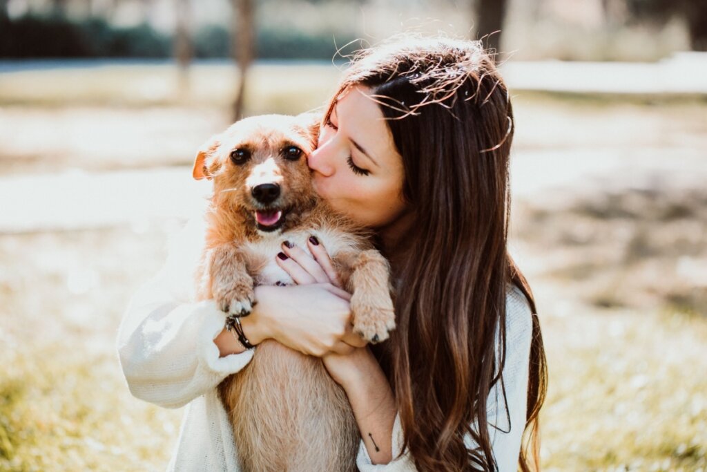 Mulher com cabelo solto, usando camisa de manga longa branca abraçada e beijando cachorro com pelagem bege ao ar livre