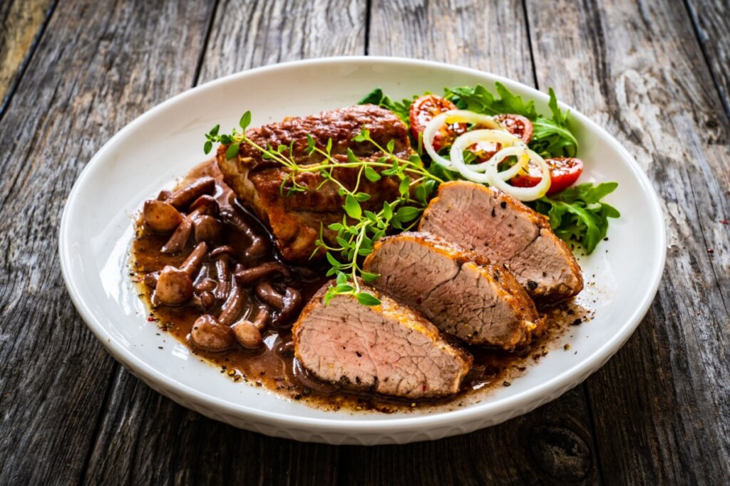 Picanha suína com molho de shimeji servida em prato branco com salada em cima de mesa de madeira