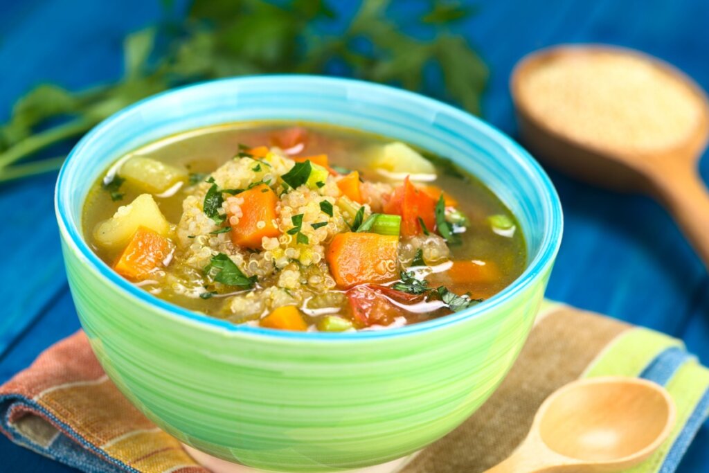 Sopa de quinoa com legumes servido em tigela de cerâmica verde em cima pano de prato listrado em cima de mesa de madeira azul