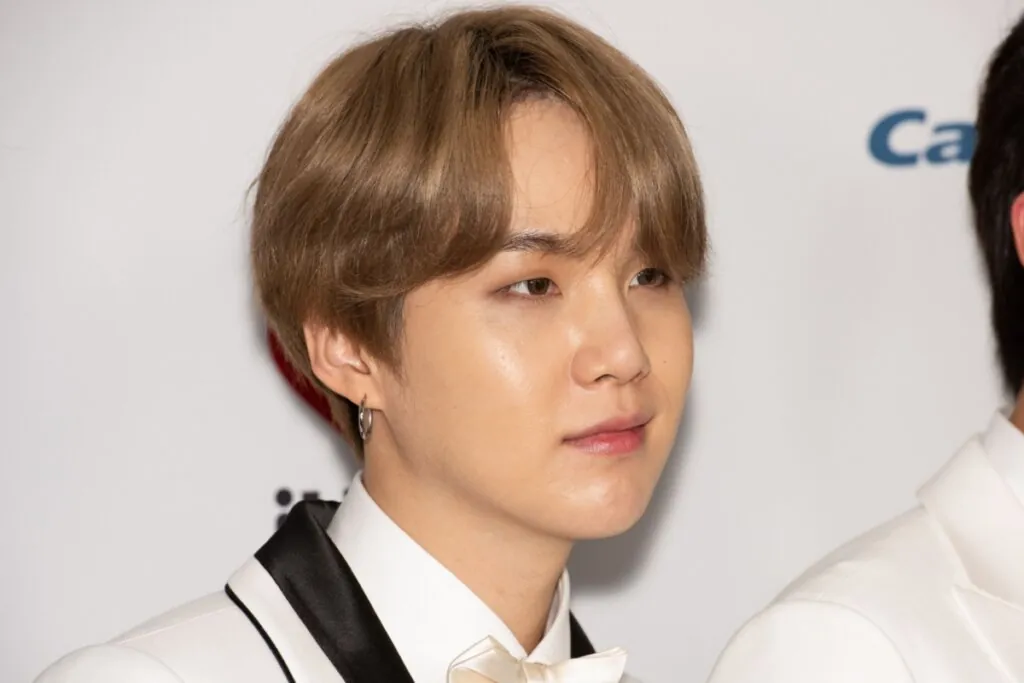 Homem com cabelo castanho claro partido ao meio com franja, usando camisa branca  e terno bege com detalhes em preto e gravata borboleta bege