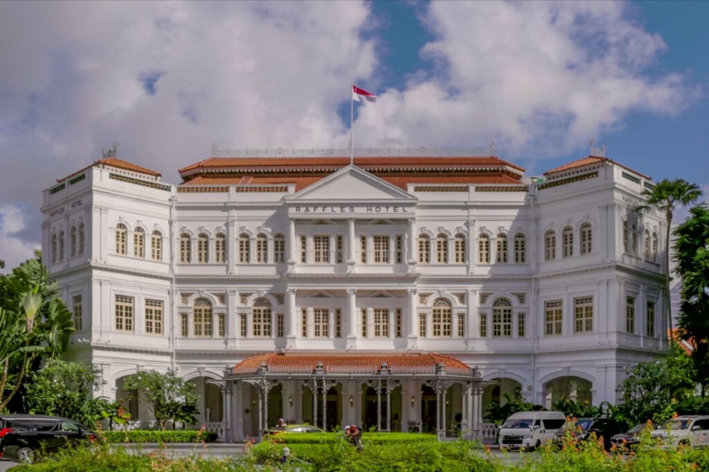 Fachada do Hotel Raffles com céu azul com nuvens acima e jardim verde na frente