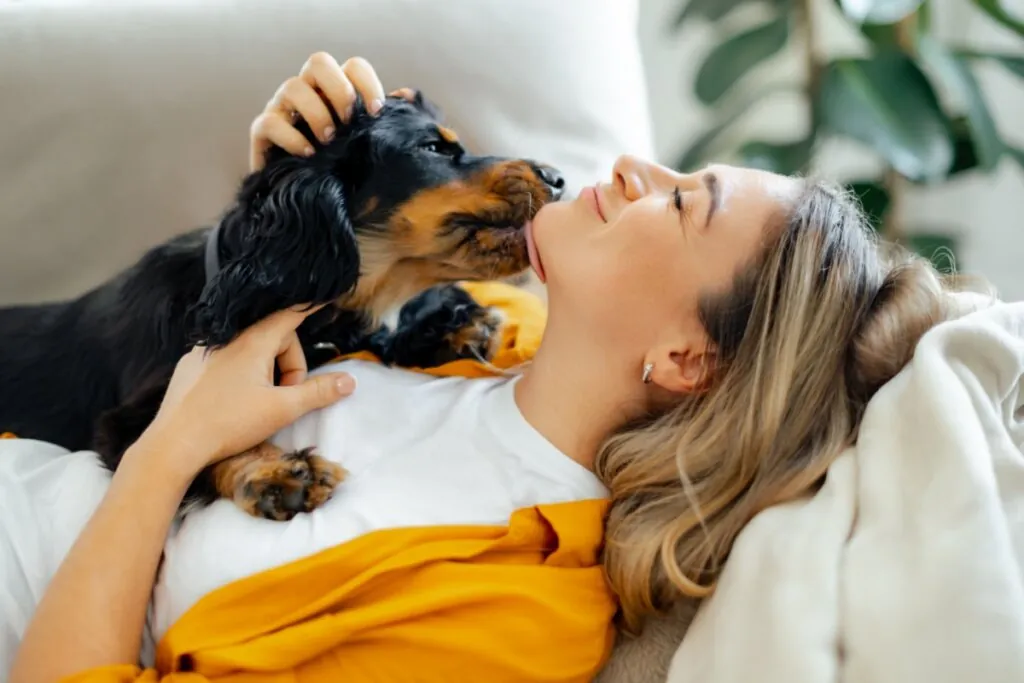 Mulher deitada com uma cachorra lambendo o seu rosto