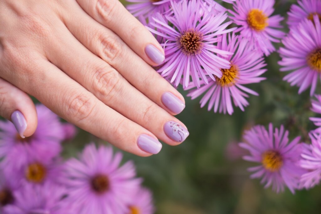 Mãos com unhas longas pintadas com esmalte lilás-claro. Ao fundo, flores lilás