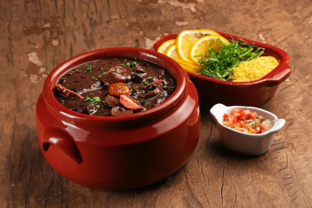 Recipiente de vidro marrom com feijoada e linguiça em cima de uma mesa de madeira com dois recipientes contendo laranja e tomate