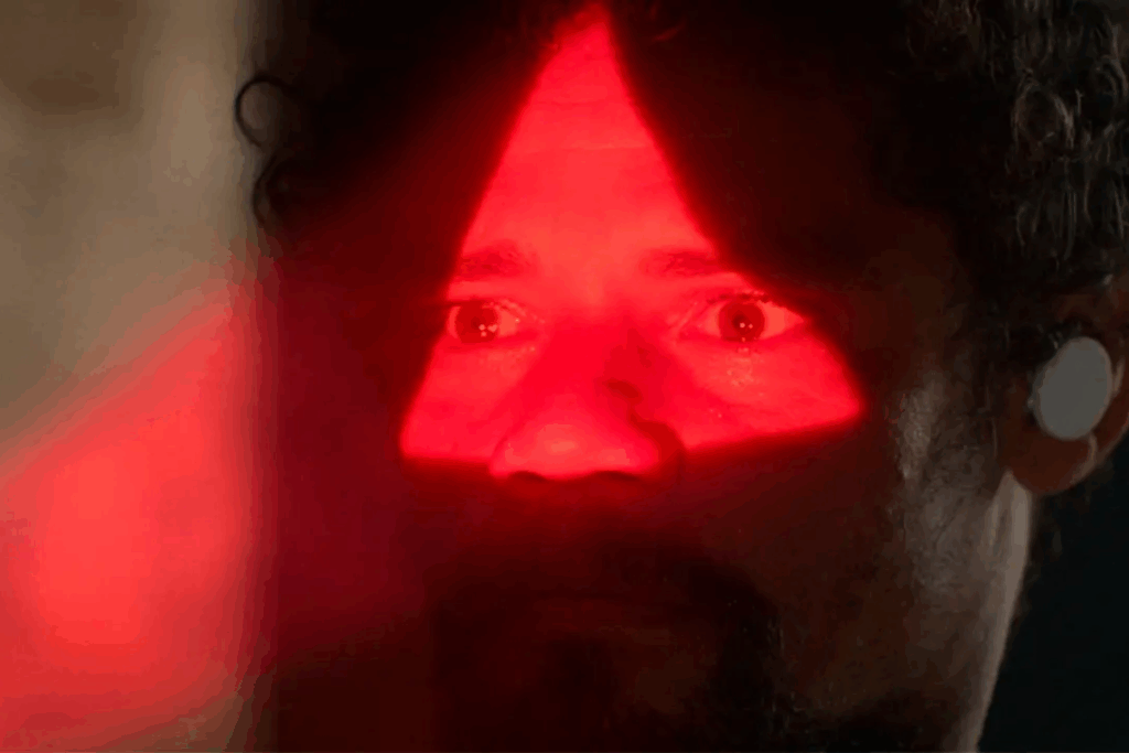 Em plano fechado, homem de cabelos cacheados e barba tem a região dos olhos e do nariz iluminada por um feixe de luz vermelha em formato triangular. Ele veste um fone de ouvido branco na orelha esquerda.