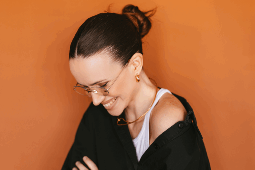 Mulher sorrindo, usando camisa preta e um coque no cabelo, em frente a um fundo laranja