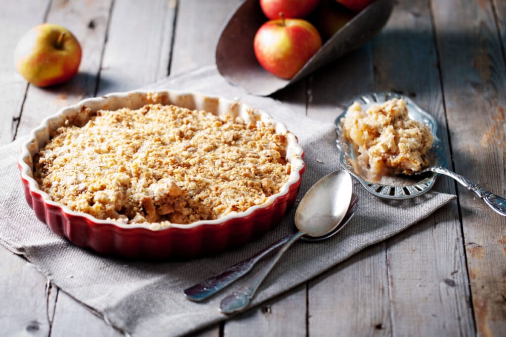 Forma vermelha com crumble de maçã em cima de uma mesa de madeira e ao lado de maçãs