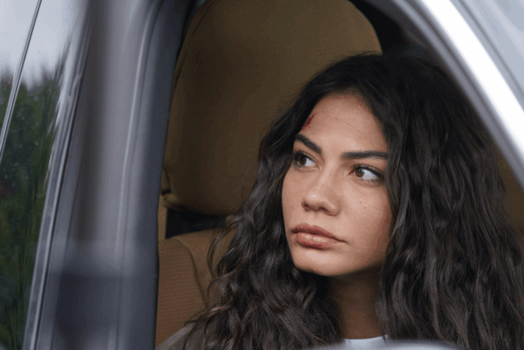 Mulher com o cabelo longo preto, machucado na testa sentada dentro de um carro olhando para fora