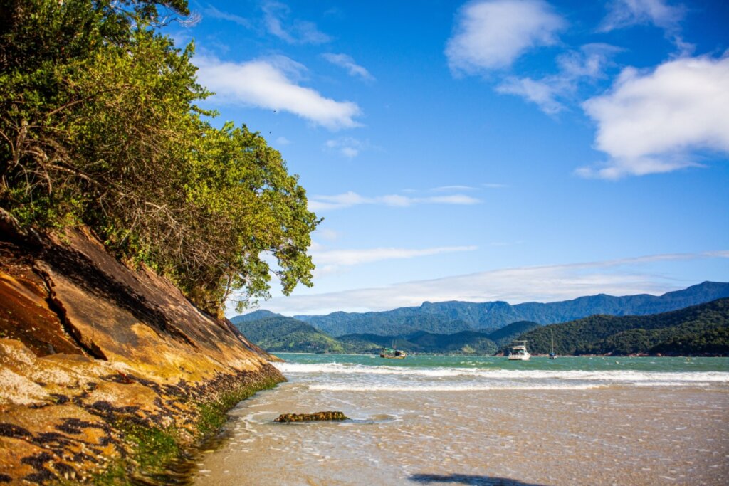Vista da Praia do Láraro com encosta com árvores, mar azul e montanhas ao fundo